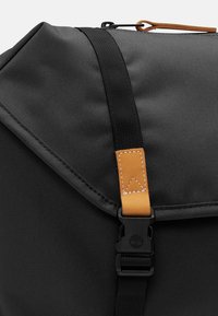 Zwarte rugzak met een klep die is beveiligd met een zwarte plastic gesp en een bruine leren band detail op een witte achtergrond.