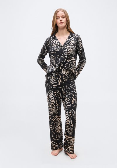 Γυναίκα μοντέλο που στέκεται ξυπόλητη φορώντας μαύρο σετ πιτζάμας με μπεζ μοτίβο ρίγες τίγρης, μακριά μανίκια και πάνω μέρος με κουμπιά.