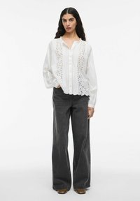 Frau mit langen dunklen Haaren, die eine weiße bestickte Bluse mit langen Ärmeln, schwarze hochtaillierte weit geschnittene Jeans und braune Schuhe trägt und steht.