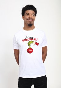 Bílý tričko s potiskem ruky Grinche držící červenou ozdobu. Text zní „Veselé GRINCHMAS!“ v černé a červené.