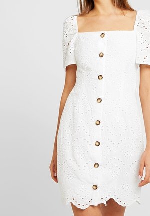 Biała bawełniana sukienka z krótkimi bufkami, kwadratowym dekoltem i dekoracyjnym wzorem ażurowym. Z przodu ozdobiona złotymi guzikami i wykończona falbanką na dole.