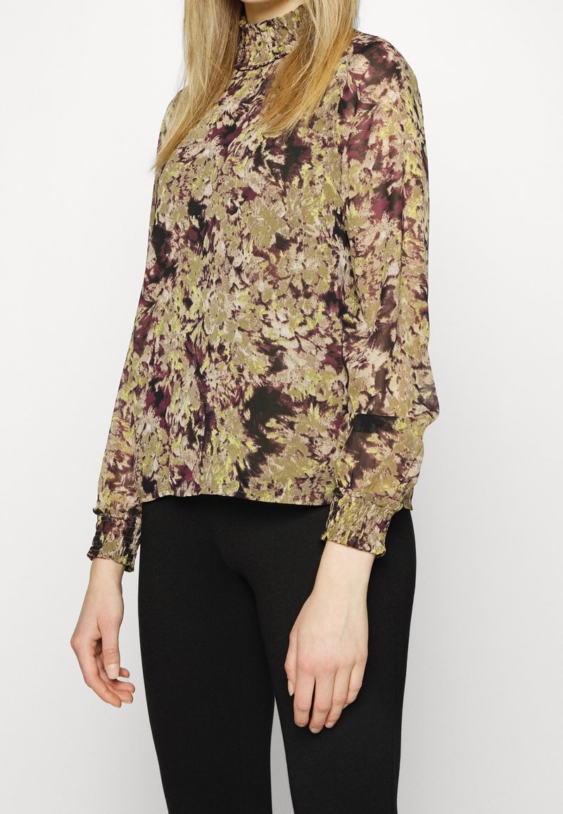 Femme portant un chemisier à manches longues avec col montant et motif floral dans des tons verts, marron et violet, associé à un pantalon noir, debout devant un fond uni.