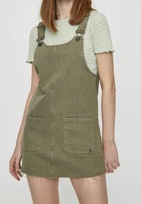 Robe salopette en denim kaki avec bretelles ajustables, deux poches avant et ourlet effiloché. Associée à un t-shirt à manches courtes rayé vert et blanc.