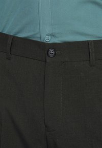 Dunkelgrüne Hose mit glatter Textur, ausgestattet mit einem Knopfverschluss, seitlichen Taschen und dezenten Nähten. Kombiniert mit einem hellblauen Hemd.