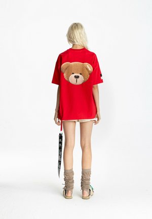 Mujer rubia con camiseta roja extragrande con un gran dibujo de un osito en la espalda, pantalones cortos beige, calentadores de piernas y zapatos, de pie mirando hacia atrás.