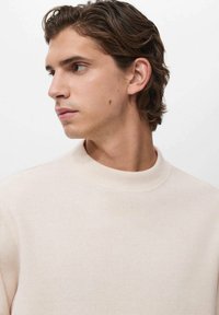 Suéter color beige claro con un cuello alto, textura acanalada y una silueta ajustada, llevado por una persona con cabello de longitud media y un fondo neutro.