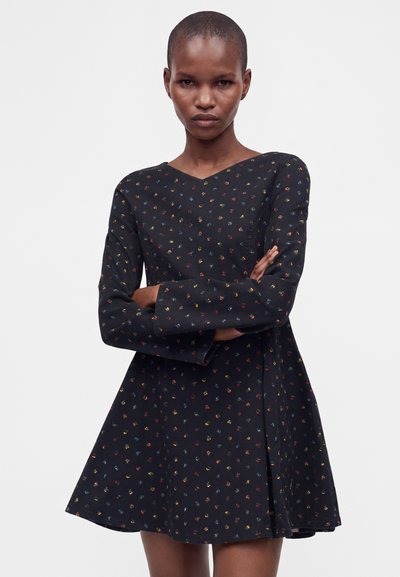Femme aux cheveux courts portant une robe noire à manches longues avec de petits motifs floraux colorés, bras croisés, regardant devant elle.
