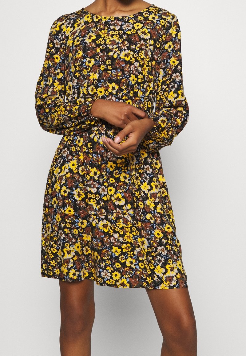 Robe fleurie avec un fond noir orné de fleurs jaunes, brunes et bleues. Manches longues et encolure ronde, avec une taille cintrée.