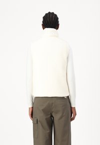 Person mit geflochtenem Haar, der eine weiße Fleece-Weste und ein Langarmoberteil trägt, kombiniert mit olivgrünen Cargo-Hosen, von hinten gezeigt.