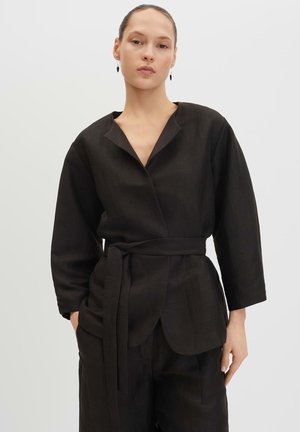 Frau mit zurückgegelten Haaren, die eine schwarze Jacke mit Gürtel und passende Hose trägt, steht vor hellem Hintergrund mit den Händen in den Taschen.