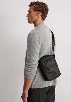 Homme portant une chemise grise à manches longues et un sac en cuir noir à bandoulière ajustable, se tenant de profil sur un fond uni.
