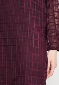 Burgundské šaty se strukturovanou tkaninou s mřížkovým vzorem, dlouhými rukávy a poloprůhlednou vrstvou. Pokožka je viditelná v blízkosti ruky.