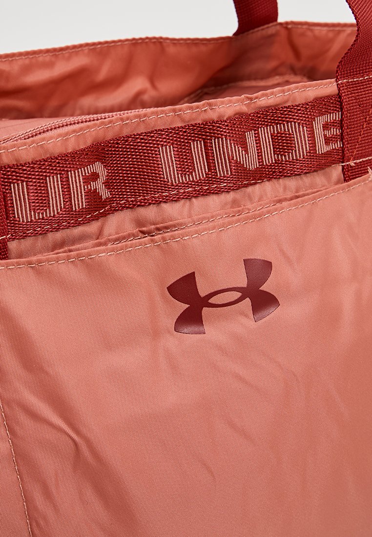 Bolso tote de color rosa claro, hecho de un tejido duradero, que cuenta con una correa estampada con el texto "UNDER" y un logo de color rosa oscuro en el lateral.