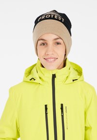Veste imperméable jaune avec des zips noirs et un logo, associée à un bonnet tricoté beige et marine. Jeune souriant avec des cheveux bruns.