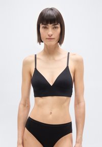 Γυναίκα με κοντά σκουρόχρωμα μαλλιά που φοράει ένα μαύρο μπραλέτο και μαύρο εσώρουχο, στέκεται μπροστά σε ένα απλό φωτεινό φόντο, κοιτάζοντας προς τα εμπρός.