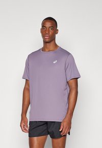 Lilla sport T-shirt med rund halsudskæring, korte ærmer, glat tekstur og et lille hvidt logo foran. Sorte shorts nedenunder.