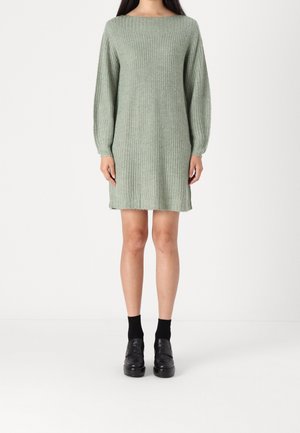 Kobieta w miętowozielonej, dzianinowej sukience z długim rękawem, czarnych skarpetkach i czarnych loafersach na platformie, stojąca na tle białego tła.