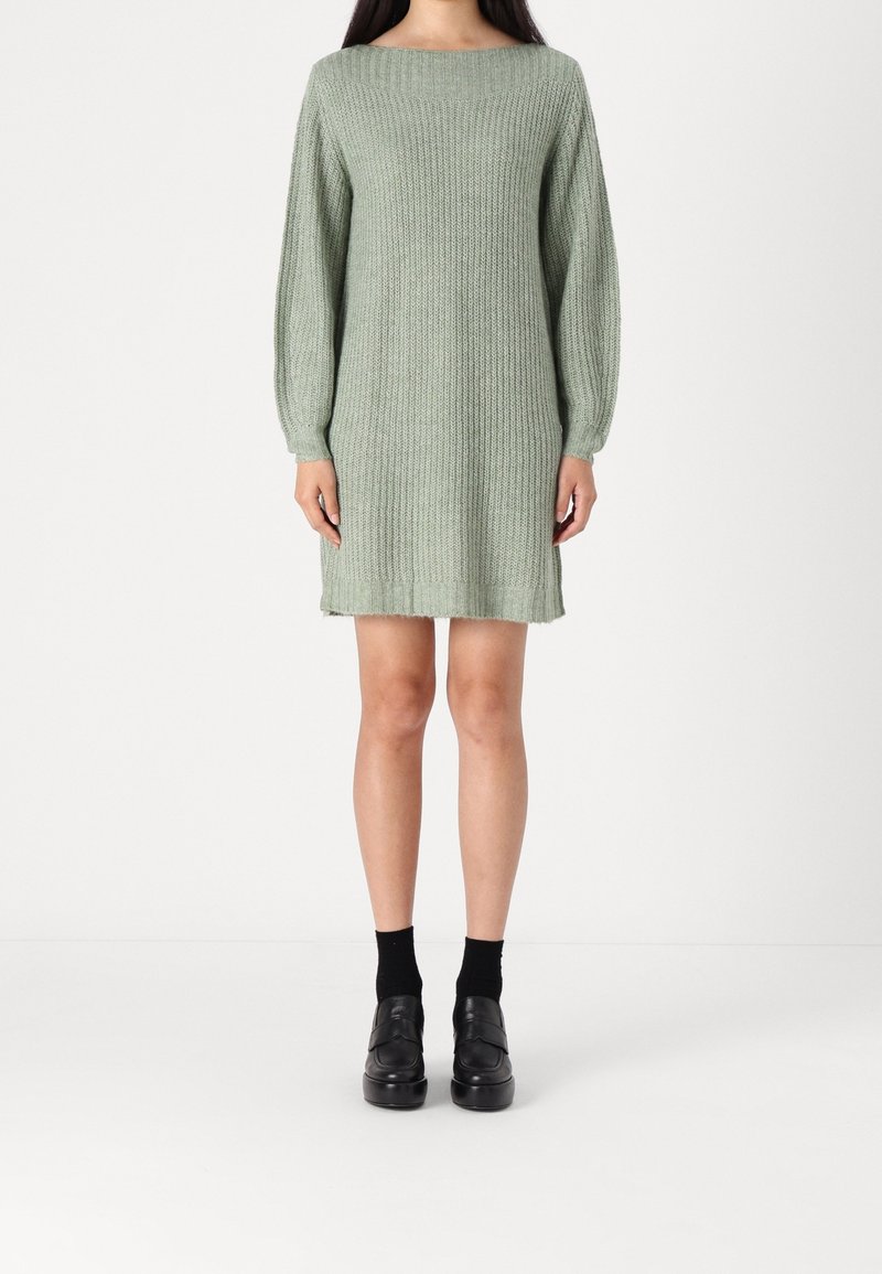Femme portant une robe longue à manches longues en tricot vert sauge, des chaussettes noires et des mocassins à plateforme noirs, debout devant un fond blanc.