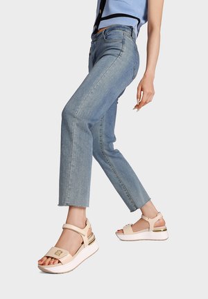 Donna con jeans corti azzurri chiari e sandali beige con zeppa e cinturini alla caviglia su sfondo semplice.
