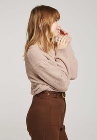 Un pull rose, texturé, avec des manches longues, porté sur un pantalon marron taille haute avec une poche latérale et une ceinture avec une boucle ronde.