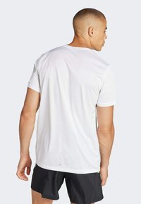 Camiseta atlética blanca de manga corta con textura suave, cuello redondo y costuras mínimas, combinada con pantalones cortos negros, mostrando un ajuste casual.
