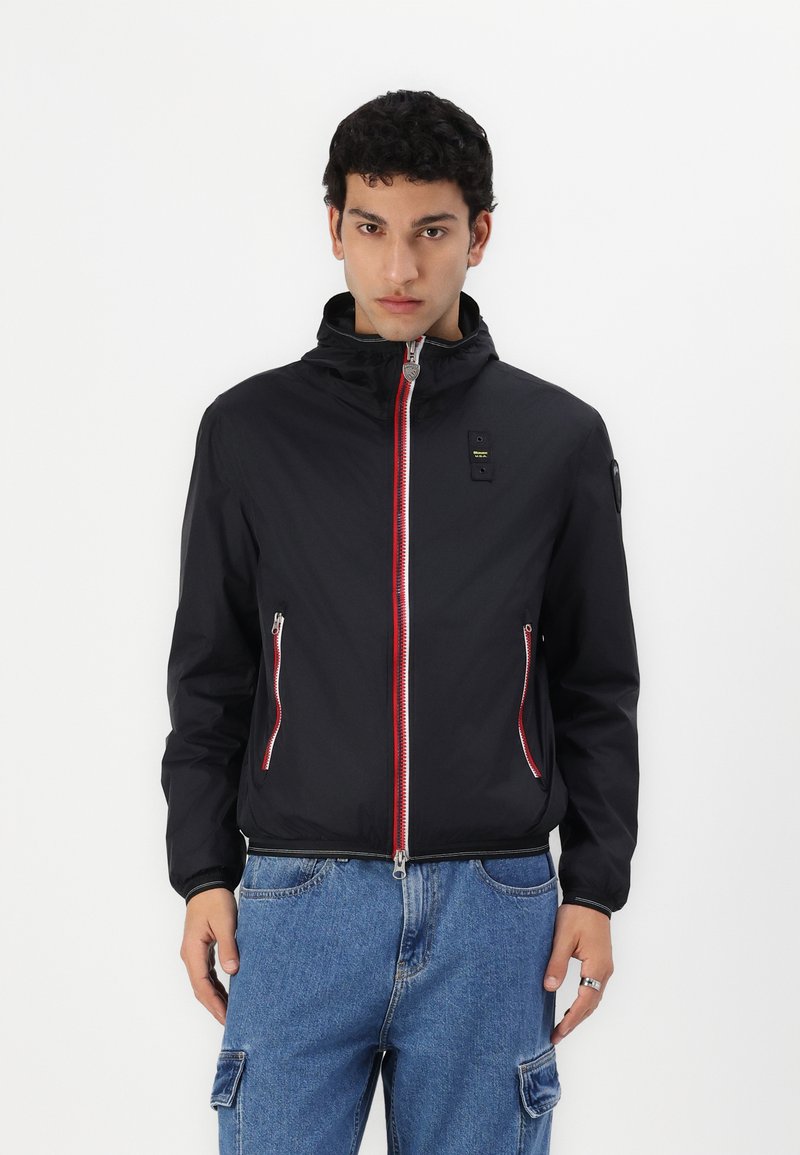 Chaqueta ligera negra con acentos en cremallera rojos y blancos, dos bolsillos laterales y cuello alzado, hecha de un material liso y resistente al agua.