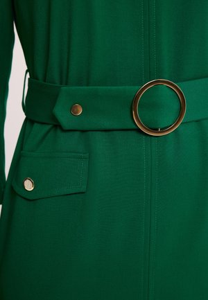 Zielona sukienka z tkaniny z szerokim paskiem ozdobionym dużą, okrągłą złotą klamrą oraz klapką kieszeni zapinaną na złoty guzik.