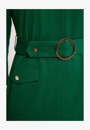 Zielona sukienka z tkaniny z szerokim paskiem ozdobionym dużą, okrągłą złotą klamrą oraz klapką kieszeni zapinaną na złoty guzik.