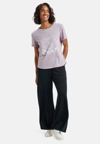 Camiseta de manga corta lila con un diseño gráfico blanco que presenta flores y un estallido de sol, combinada con pantalones negros holgados y zapatillas blancas.