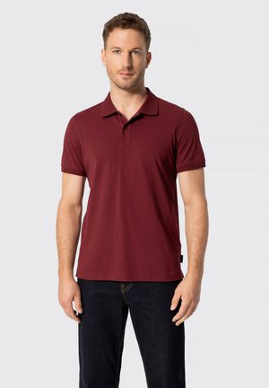 Hombre con cabello corto y rizado, vestido con una camisa polo burdeos de manga corta y vaqueros oscuros, de pie frente a un fondo claro y liso.