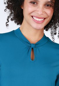 Türkise Bluse mit kurzen Ärmeln, Schlüssel-Ausschnitt am Hals und weicher, glatter Stoffstruktur.