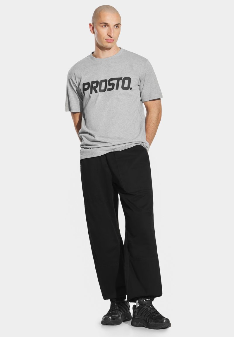 Szara bawełniana koszulka z czarnym napisem "PROSTO.", w połączeniu z luźnymi czarnymi spodniami i masywnymi czarnymi sneakersami, na białym tle.