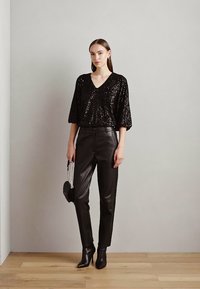 Schwarzes Pailletten-Oberteil mit V-Ausschnitt, kombiniert mit eng anliegenden schwarzen Kunstlederhosen. Das Modell trägt schwarze Stiefeletten mit Absatz und hält eine kleine Handtasche.