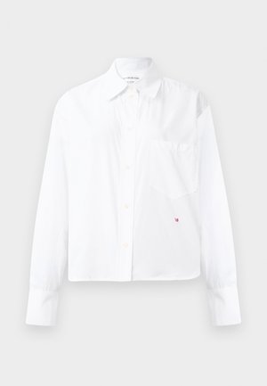 Camicia bianca a maniche lunghe con bottoni, colletto a punta e taschino sul petto, caratterizzata da un piccolo ricamo rosso "VB" vicino all'orlo.