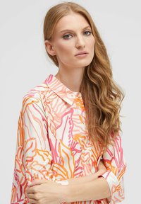 Junge Frau mit langen, welligen Haaren, die ein button-up Hemd mit pinken, orangefarbenen und weißen Mustern trägt, die Arme vor einem schlichten Hintergrund verschränkt.