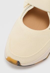 Lys beige semsket skinnsko med et elastisk bånd og rund tå. Har en lys brun innvendig og en klar gummisåle med en svak fargegradient.