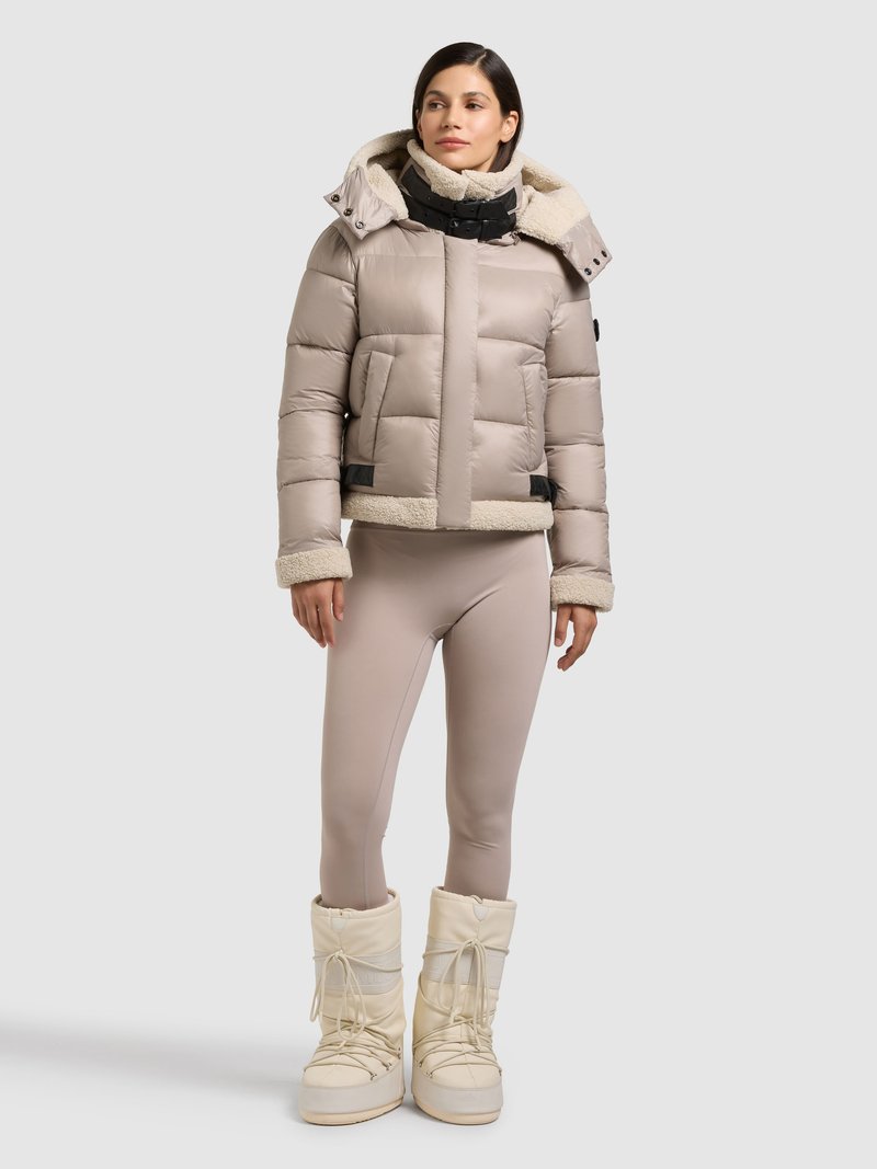 Giacca imbottita beige con colletto in shearling, due tasche frontali e chiusura nera. Abbinata a leggings color nudo e stivali invernali bianchi.