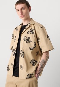 Beige rövid ujjú ing, pamut anyagból, fekete virágmintákkal, gombos záródással és mellzsebből áll. Fekete inggel párosítva alatta.