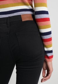 Jeans de mezclilla negra con una textura suave, que cuentan con una etiqueta de cuero marrón en la cintura. Un suéter de rayas colorido es parcialmente visible en las mangas.