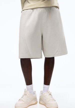 Osoba ubrana w jasnobeżowe spodenki do kolan, białe skarpetki i kremowe trampki, stojąca na białym tle.