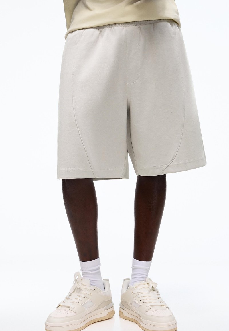 Persoană purtând pantaloni scurți bej deschis până la genunchi, șosete albe și adidași culoarea alb murdar, stând în fața unui fundal alb.