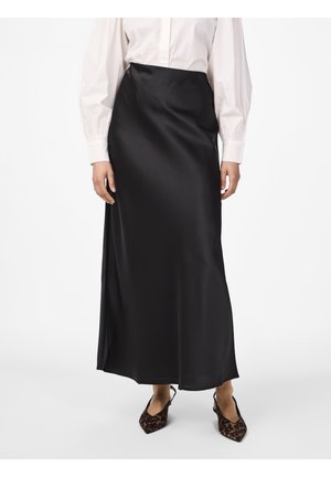 Kobieta ubrana w czarną satynową długą spódnicę, białą bluzkę z długim rękawem oraz cieliste sandały typu slingback w zwierzęcy wzór, stojąca na białym tle.