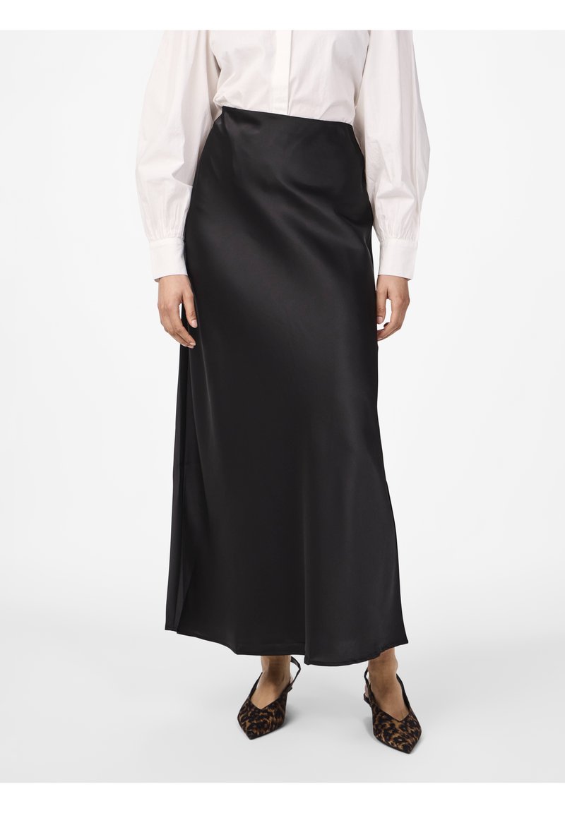 Femme portant une jupe maxi en satin noire, un chemisier blanc à manches longues et des chaussures slingback à bout pointu imprimé léopard, debout sur fond blanc.