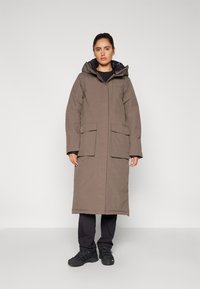 Long brown parka with a hood, featuring two front pockets, a smooth texture, and a slightly loose fit. Worn over dark pants and shoes.