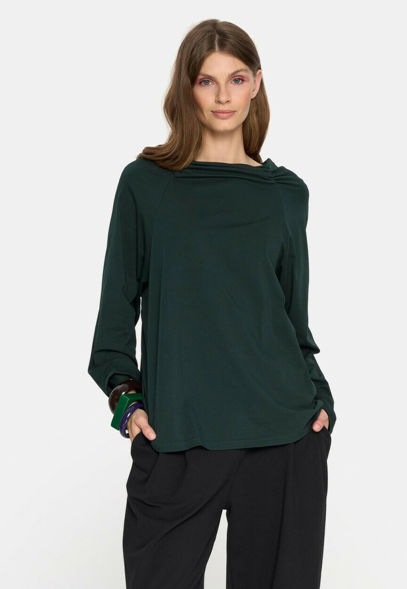 Kobieta w ciemnozielonej bluzce z długim rękawem i luźnym dekoltem, czarnych spodniach oraz kolorowych bransoletkach, stojąca z rękami w kieszeniach.