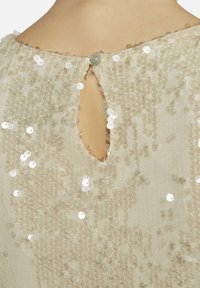 Beiges Paillettentop mit einem Schlüsselaugen-Rücken und einer einzelnen Knopfverschluss sowie einer transparenten Textur, die mit glänzenden, reflektierenden Pailletten verziert ist.