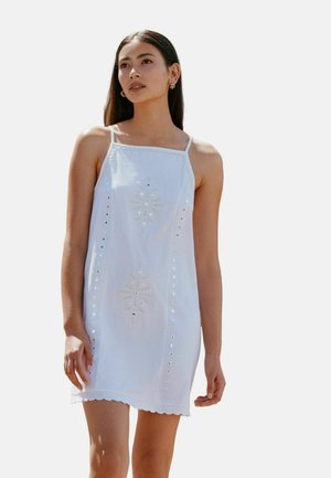 Junge Frau mit langem, dunklem Haar, die ein ärmelloses weißes Kleid mit bestickten Blumenmustern und ausgeschnittenem Saum trägt.