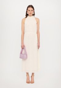 White, textured maxi dress with a halter neck and cinched waist. Holding a small, lavender bucket bag. Strappy nude heels complete the look.