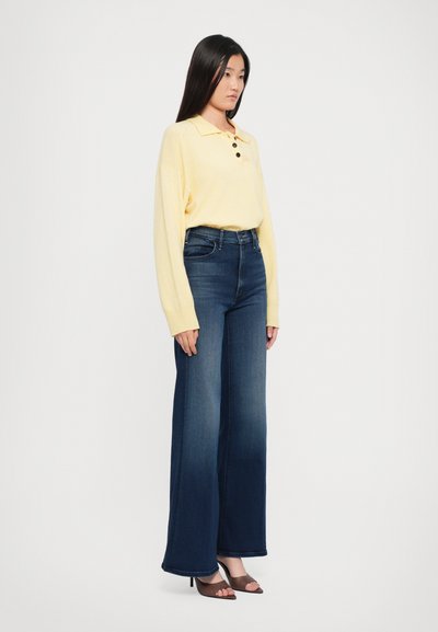 Polo amarillo de manga larga con cuello y tres botones, combinado con vaqueros de pierna ancha azul oscuro y sandalias de tacón marrones.