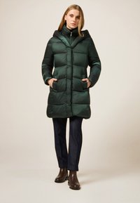 Mørkegrøn dunjakke med hætte, quiltet design, forlommer og lynlåslukning. Kombineret med mørke jeans og brune ankelstøvler.